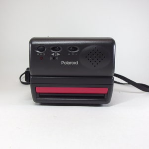 Peut inclure: Un appareil photo Polaroid noir avec une bande rouge sur le devant. L'appareil photo est doté d'un flash intégré et d'une sangle.