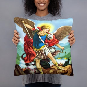 May include: A throw pillow featuring a colourful painting of the Archangel Michael, a winged figure in armour, standing on a dragon with a sword in his hand and scales in the other.