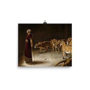 May include: A framed print depicting a biblical scene. A man in a red robe stands before a group of lions in a dark, stone chamber. The lions are a light brown color, and the man is looking up.