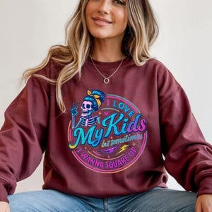 May include: Burgundy sweatshirt with a colourful graphic. The graphic features a skeleton with a blue bow and the text "I Love My Kids but sometimes I wanna square up". The sweatshirt has long sleeves and a crew neck.