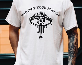 Puede incluir: Camiseta blanca con el texto "PROTECT YOUR ENERGY" sobre un diseño de ojo negro. El ojo tiene detalles intrincados y una forma de media luna debajo. Prenda de vestir informal.