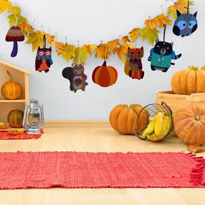 Puede incluir: Una exhibici&oacute;n con tem&aacute;tica oto&ntilde;al que presenta una guirnalda de adornos de animales de fieltro, calabazas y hojas de oto&ntilde;o. Los adornos incluyen un b&uacute;ho, un oso, una ardilla y una calabaza. Una alfombra roja y una linterna completan la escena.