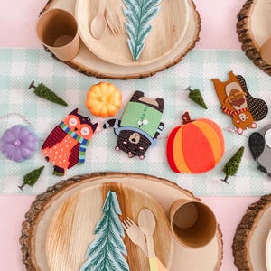 Puede incluir: Una mesa festiva con platos, tazas y cubiertos de madera. Recortes decorativos de fieltro de un b&uacute;ho, un oso, una ardilla y calabazas est&aacute;n ensartados. La mesa est&aacute; decorada con peque&ntilde;os &aacute;rboles verdes y un mantel a cuadros azul claro y blanco.
