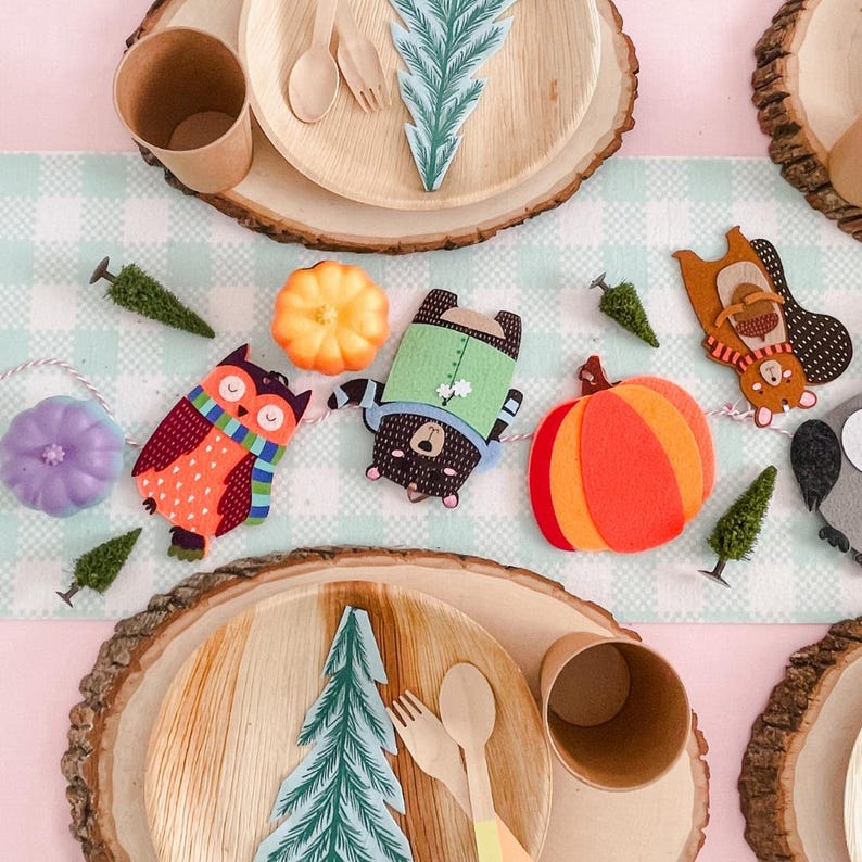 Puede incluir: Una mesa festiva con platos, tazas y cubiertos de madera. Recortes decorativos de fieltro de un b&uacute;ho, un oso, una ardilla y calabazas est&aacute;n ensartados. La mesa est&aacute; decorada con peque&ntilde;os &aacute;rboles verdes y un mantel a cuadros azul claro y blanco.