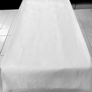 May include: A white table runner made of a soft, smooth fabric. The runner is laying flat on a white tiled surface.