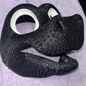 May include: A black, textured mask with white eye surrounds. The mask has a detailed, woven appearance and a curved shape, possibly for a costume or cosplay. The design includes a snout-like projection and is likely 3D-printed.