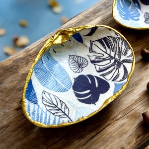 Peut inclure: Deux plats décoratifs en céramique en forme de coquillages. Les plats sont peints avec un fond blanc et un motif de feuilles bleues et noires. Les bords des plats sont bordés de peinture dorée.