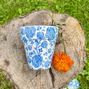 May include: A small, white ceramic flower pot with a blue floral pattern. The pot is sitting on a tree stump with an orange flower and a green stem. The background is green grass.