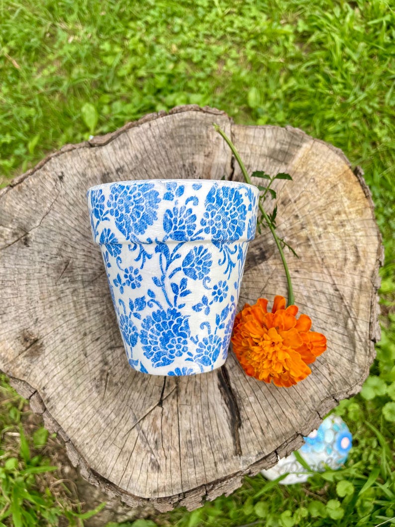 May include: A small, white ceramic flower pot with a blue floral pattern. The pot is sitting on a tree stump with an orange flower and a green stem. The background is green grass.