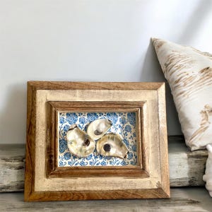 May include: A framed wall decor featuring three oyster shells on a blue and white floral patterned background. The frame is made of wood with a light brown finish.