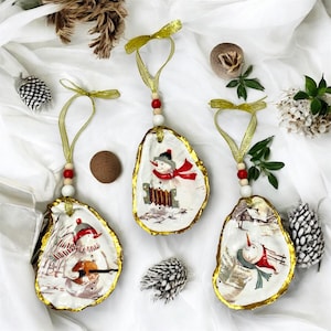 Op de afbeelding: Drie kerstversieringen van oesterschelpen. Elke schelp is met de hand beschilderd met een sneeuwmanontwerp, met een rode sjaal en een zwarte hoed. De schelpen hebben een gouden rand, een gouden lint en houten kralen om op te hangen. De achtergrond is wit.