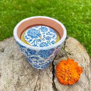 May include: A ceramic candle holder with a blue floral pattern. The candle inside is yellow, and the holder is sitting on a tree stump next to an orange flower. The candle holder has a white rim and a brown interior.