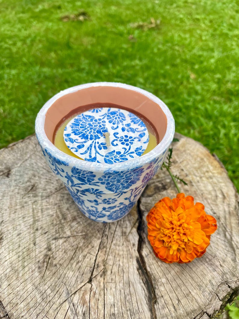May include: A ceramic candle holder with a blue floral pattern. The candle inside is yellow, and the holder is sitting on a tree stump next to an orange flower. The candle holder has a white rim and a brown interior.