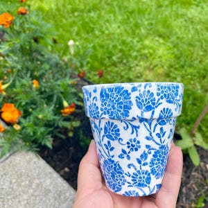 May include: A small, white flower pot with a blue floral pattern. The pot is held in a hand, with a blurred background of green grass and orange flowers.