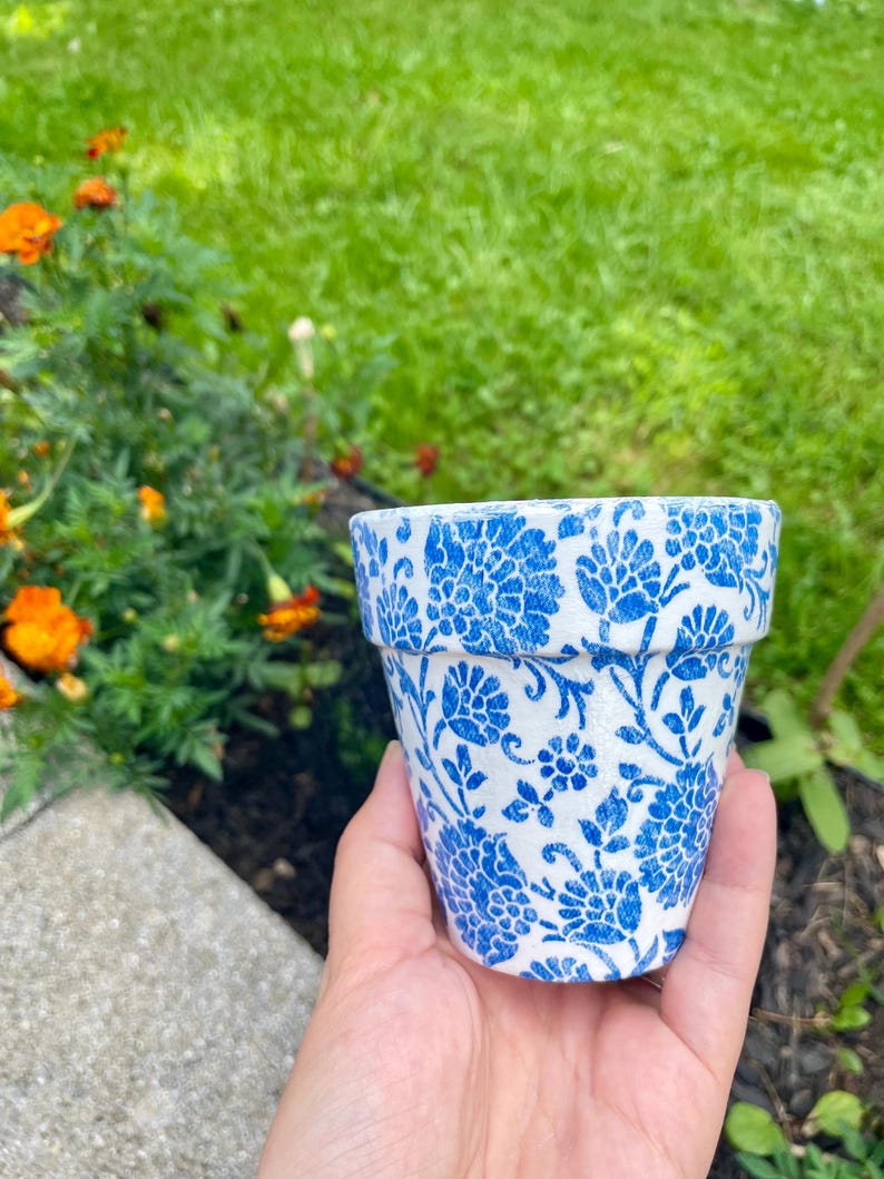 May include: A small, white flower pot with a blue floral pattern. The pot is held in a hand, with a blurred background of green grass and orange flowers.