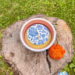 May include: A round candle in a white and brown container with a blue floral design on top. An orange flower rests next to the candle on a tree stump, with green grass in the background.