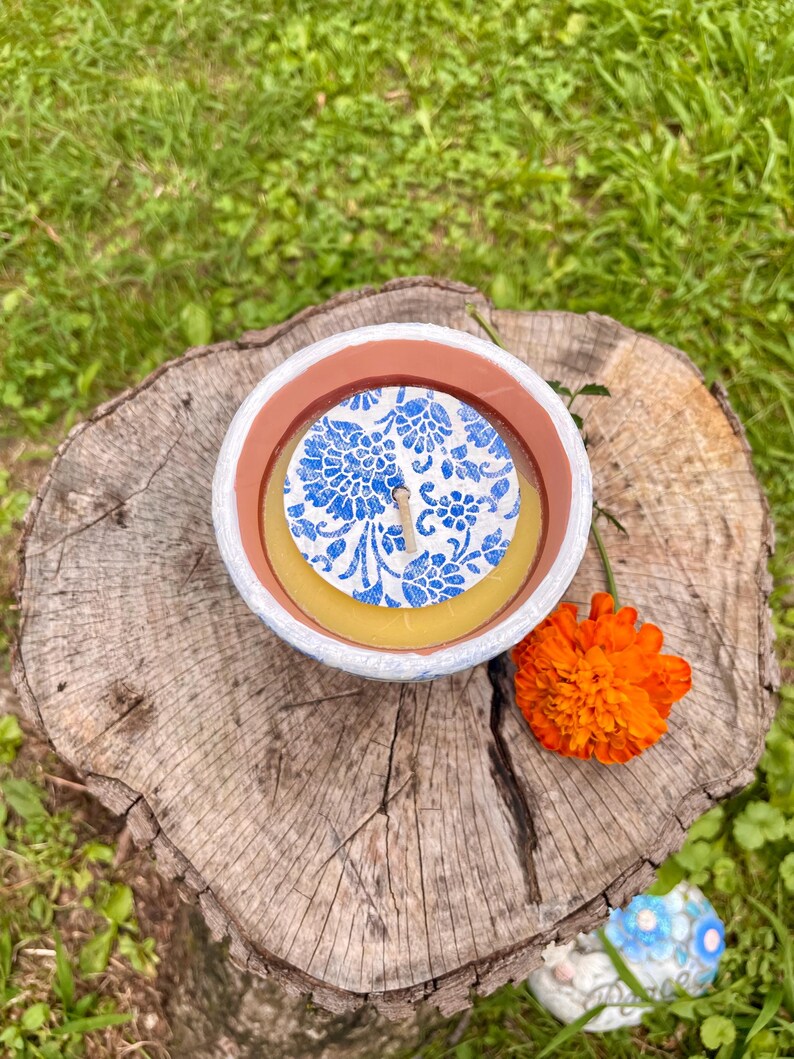 May include: A round candle in a white and brown container with a blue floral design on top. An orange flower rests next to the candle on a tree stump, with green grass in the background.