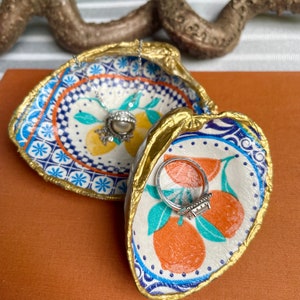 Op de afbeelding: Twee keramische oesterschelpen met een gouden rand. De ene schaal heeft een blauw en wit patroon met citroenen en een parelketting erin. De andere schaal heeft een witte achtergrond met oranje citroenen en een zilveren ring erin.