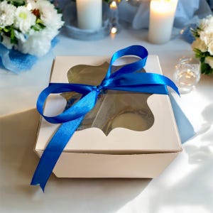 May include: A white gift box with a clear window, tied with a blue ribbon. The box is filled with small items, such as jewelry or other trinkets.