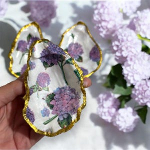 May include: Three small white oyster shell dishes with gold trim. Each dish is decorated with a purple and green floral pattern.