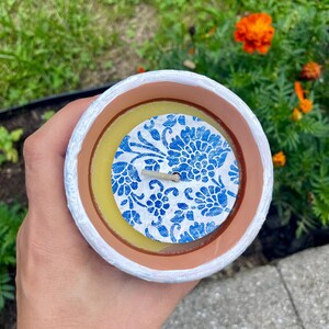 May include: A handmade candle in a white, brown, and yellow container. The candle has a blue floral design on top. The candle is held in a hand, with a green and orange floral background.