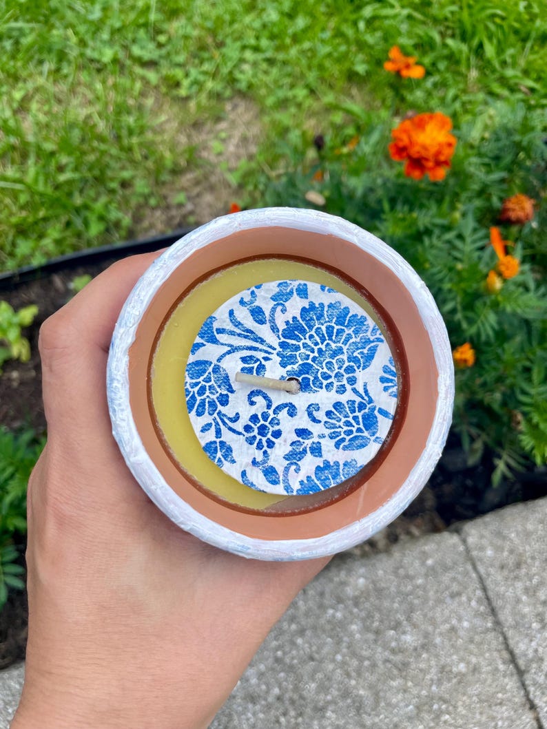May include: A handmade candle in a white, brown, and yellow container. The candle has a blue floral design on top. The candle is held in a hand, with a green and orange floral background.