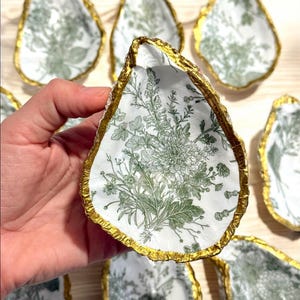 May include: A set of decorative oyster shell dishes with a white background and a green floral pattern. The edges of the shells are painted gold.