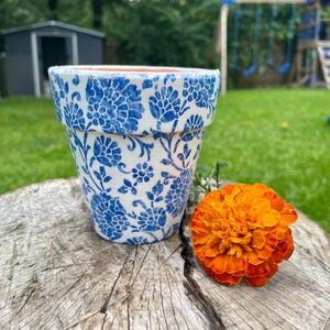 May include: A blue and white floral patterned ceramic flower pot sits on a tree stump next to an orange marigold flower. The flower pot has a classic shape and is unglazed on the inside.