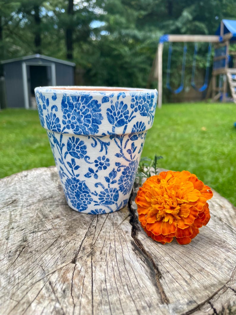 May include: A blue and white floral patterned ceramic flower pot sits on a tree stump next to an orange marigold flower. The flower pot has a classic shape and is unglazed on the inside.
