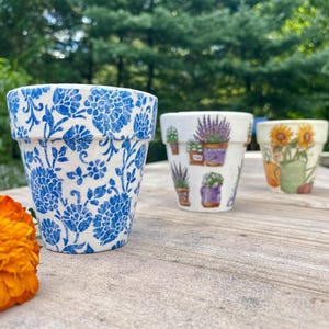 May include: Three decorative ceramic flower pots. The first pot has a blue floral pattern. The second pot has images of lavender plants. The third pot has images of sunflowers. An orange flower is in the foreground.