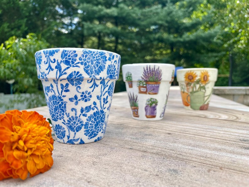 May include: Three decorative ceramic flower pots. The first pot has a blue floral pattern. The second pot has images of lavender plants. The third pot has images of sunflowers. An orange flower is in the foreground.