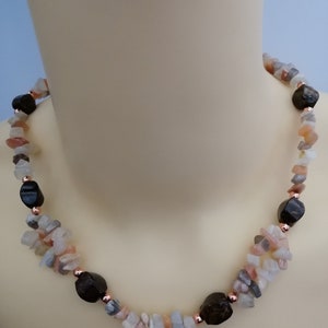 Può includere: Una collana di pietre preziose con un mix di schegge di pietra rosa chiaro, grigio e bianco, intervallate da perline sfaccettate marrone scuro. La collana è impreziosita da piccole perline color rame ed è esposta su un manichino.