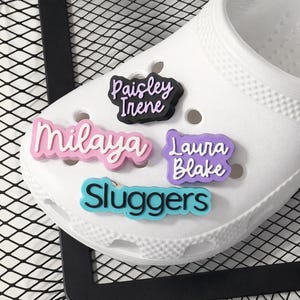 May include: White clog shoe with decorative charms. Charms include names: "Milaya" in pink, "Paisley Irene" in black, "Laura Blake" in purple, and "Sluggers" in blue. The shoe is set against a black metal grid.