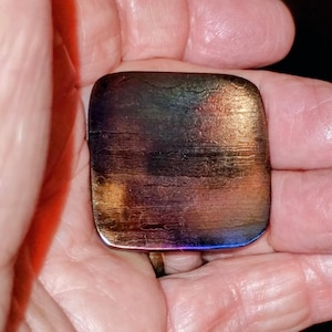 May include: A square, iridescent glass pendant with rounded corners. The colors shift from gold to purple and blue, with a textured, layered appearance. The pendant is held in a hand.