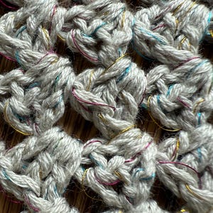 May include: Close-up of a crocheted fabric in a light gray color. The yarn has a textured appearance with subtle flecks of gold, pink, and teal. The crochet pattern creates a grid-like design, with each stitch clearly defined.