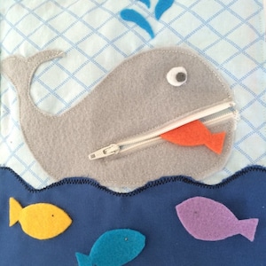 May include: A gray felt whale with a zipper mouth, a white zipper, and an orange felt fish inside the mouth. The whale is swimming in blue felt water with three felt fish: yellow, blue, and purple.