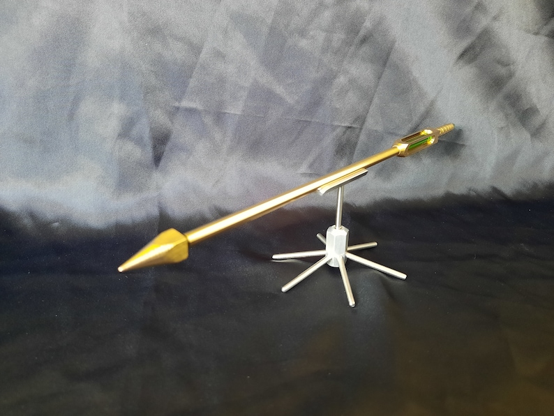May include: A gold-coloured metal arrow with a pointed tip is mounted on a silver metal stand with six legs. The arrow is on a black background.