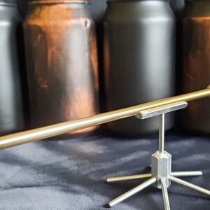 May include: A silver metal arrow with a green insert is displayed on a silver metal stand. The arrow is pointed towards the right. The stand has six legs. The arrow is in front of four cans, two black and two brown.