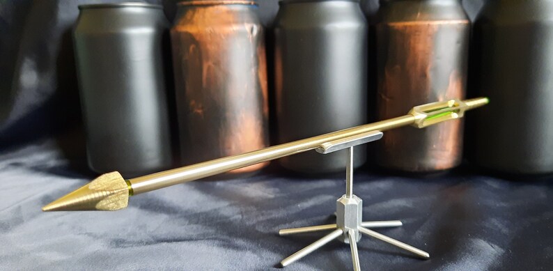 May include: A silver metal arrow with a green insert is displayed on a silver metal stand. The arrow is pointed towards the right. The stand has six legs. The arrow is in front of four cans, two black and two brown.