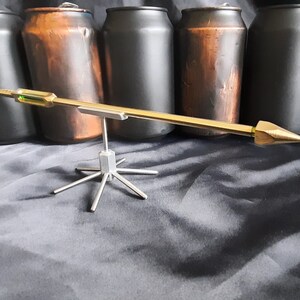 May include: A gold-coloured metal arrow with a green accent on the shaft, balanced on a silver metal stand with six legs. The arrow is resting on a black surface.