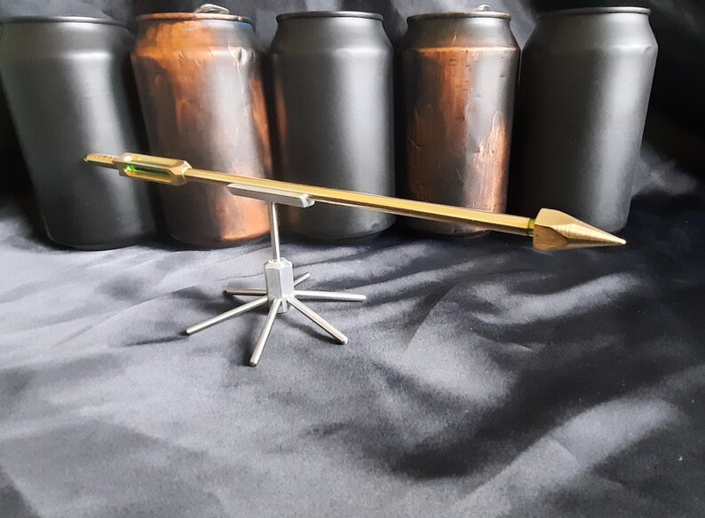 May include: A gold-coloured metal arrow with a green accent on the shaft, balanced on a silver metal stand with six legs. The arrow is resting on a black surface.