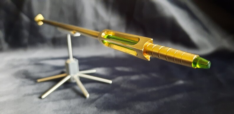 May include: A gold-coloured metal rod with a green glass section and a green tip, mounted on a silver metal stand.