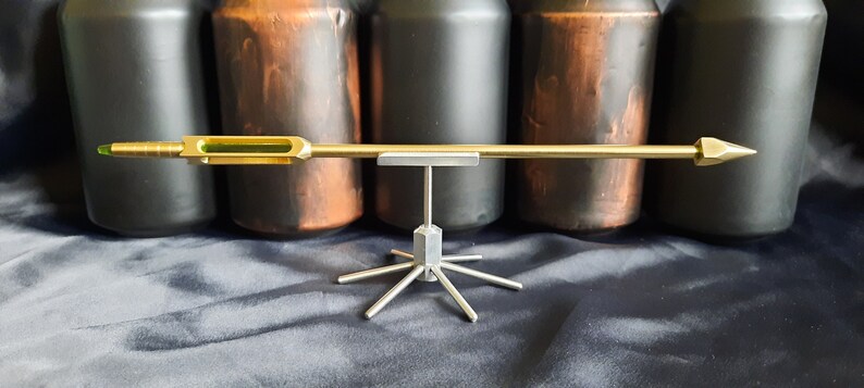 May include: A gold-coloured arrow with a green glass section and a pointed tip, displayed on a silver metal stand with multiple arms.