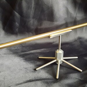 May include: A gold-coloured arrow with a green glass section is displayed on a silver metal stand. The arrow is pointed and has a sharp tip. The stand has a hexagonal base and six legs.