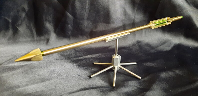 May include: A gold-coloured arrow with a green glass section is displayed on a silver metal stand. The arrow is pointed and has a sharp tip. The stand has a hexagonal base and six legs.