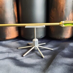 May include: A gold-coloured arrow with a green sight is displayed on a silver stand with six legs. The arrow is on a black fabric background.