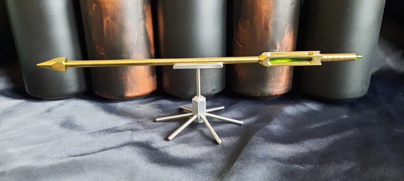 May include: A gold-coloured arrow with a green sight is displayed on a silver stand with six legs. The arrow is on a black fabric background.