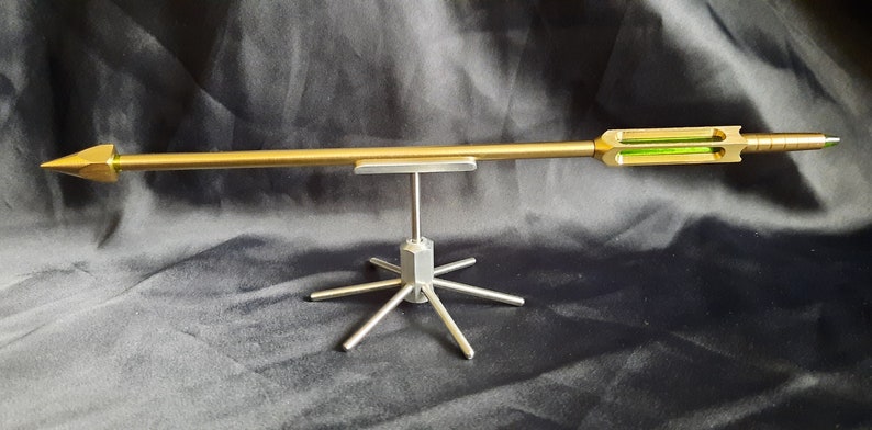 May include: A gold-coloured arrow with a green glass insert is displayed on a silver metal stand with six legs.