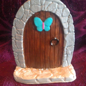 May include: A miniature stone door with a wooden door and a turquoise butterfly on the top. The door is set on a stone base.