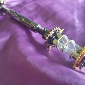 May include: A steampunk-style pen with a clear glass tube, metal gears, and a brass tip. The pen is lying on a purple fabric surface.